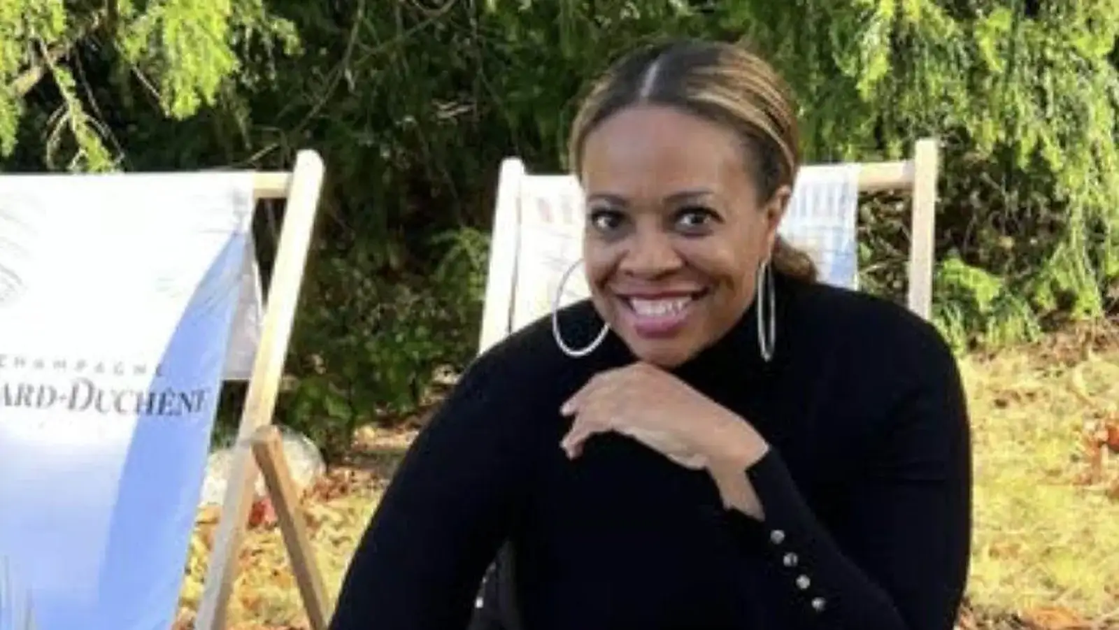 Claire B. Soares outdoors in a black turtleneck and hoop earrings, smiling beside white Champagne deck chairs under evergreen trees